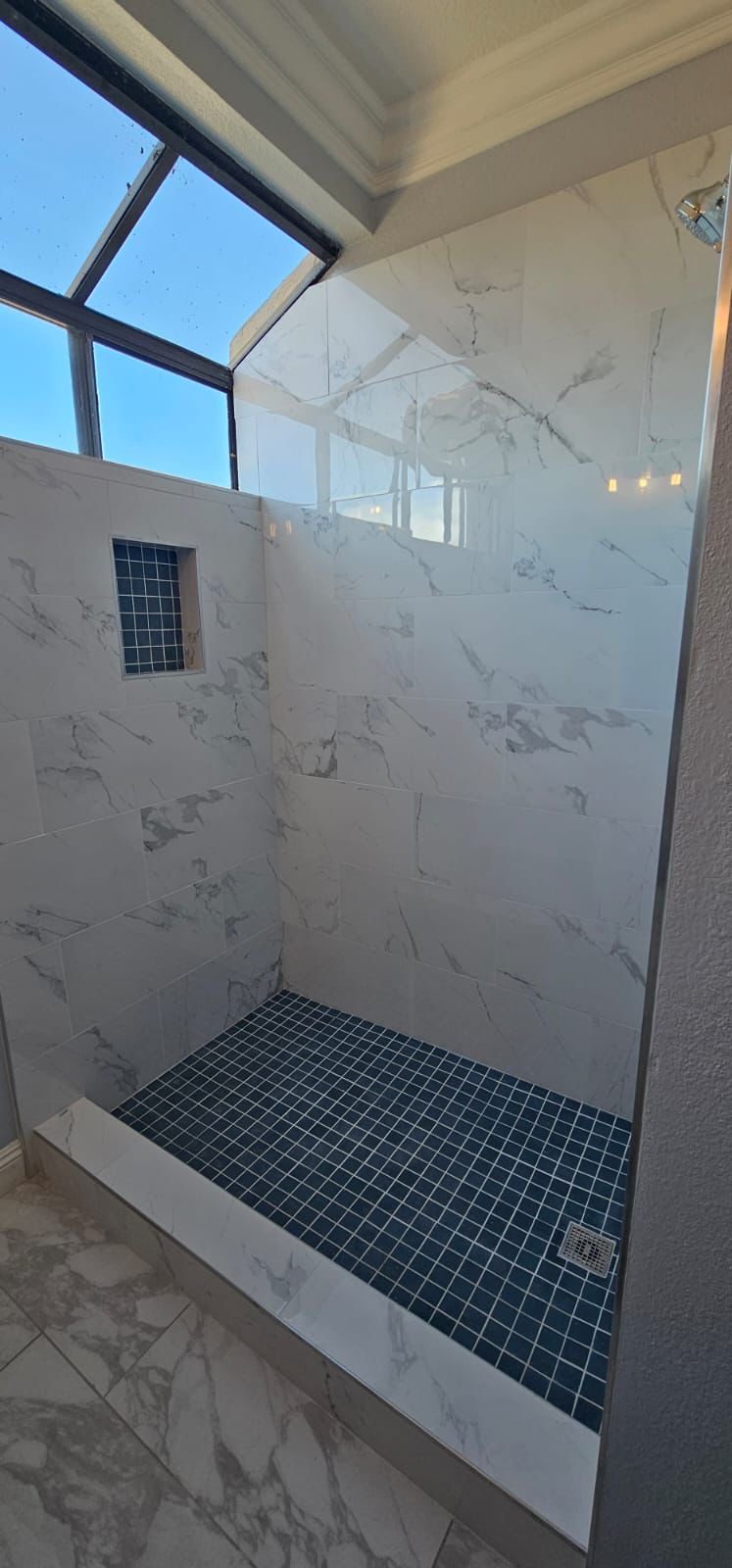 A finished walk-in shower tiled in white marble with a dark mosaic floor and skylight